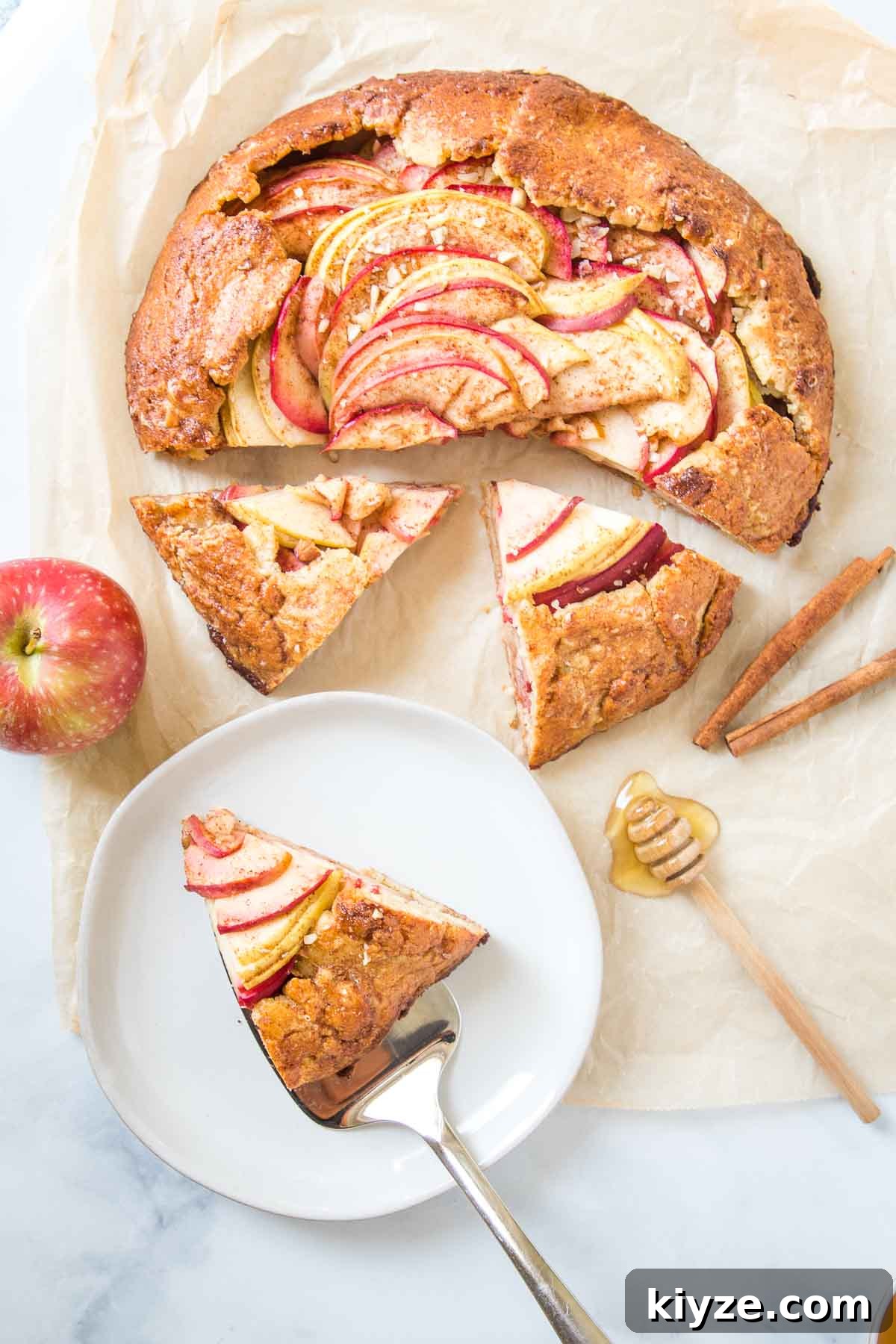 Free form apple galette, half of it sliced. One piece is on a plate. There is an apple, some cinnamon sticks and a dollop of honey on the parchment paper that the galette is also on.