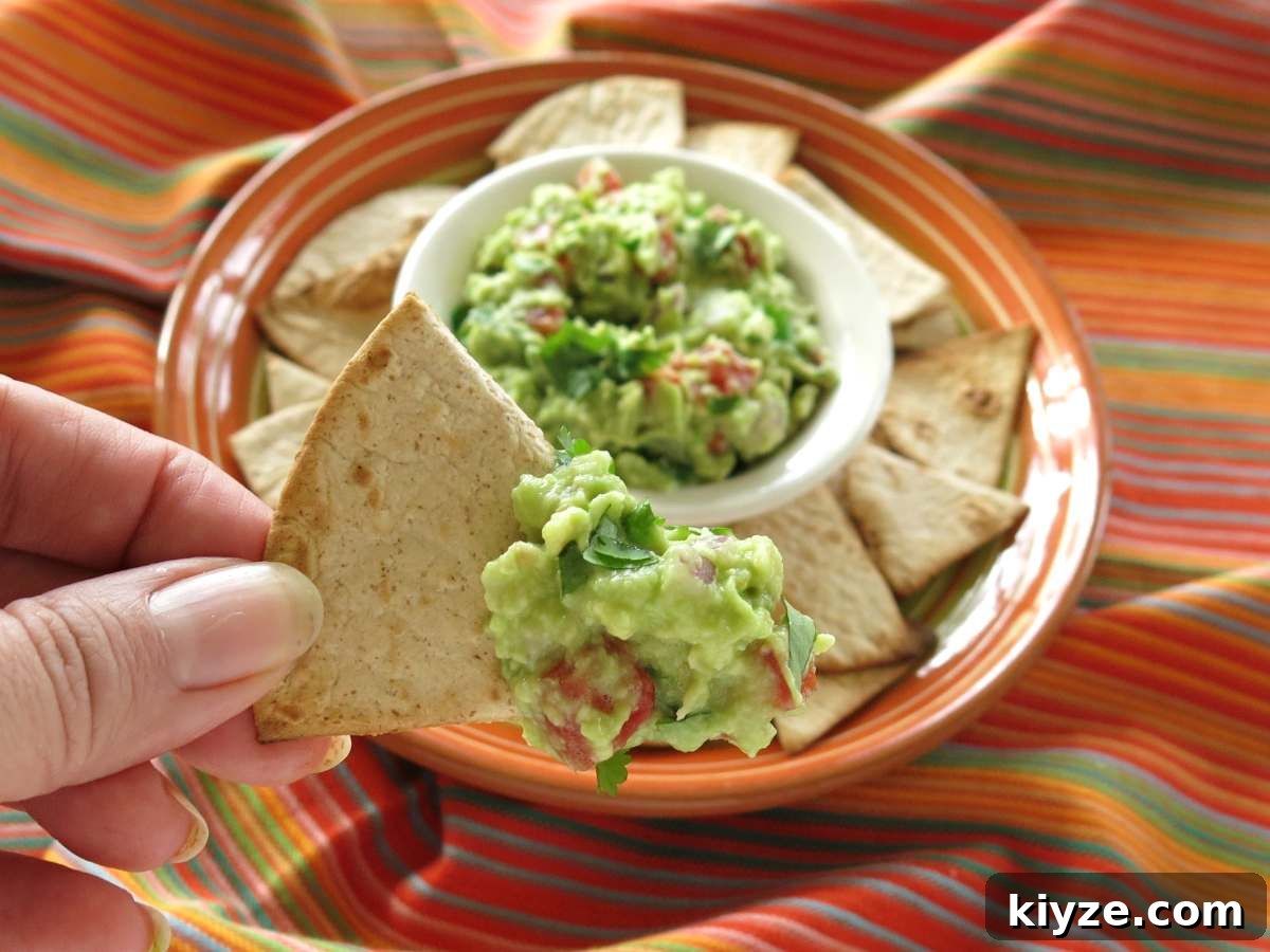 Hand holding a tortilla chip with guacamole on it with a platter of more guac and chips behind it.