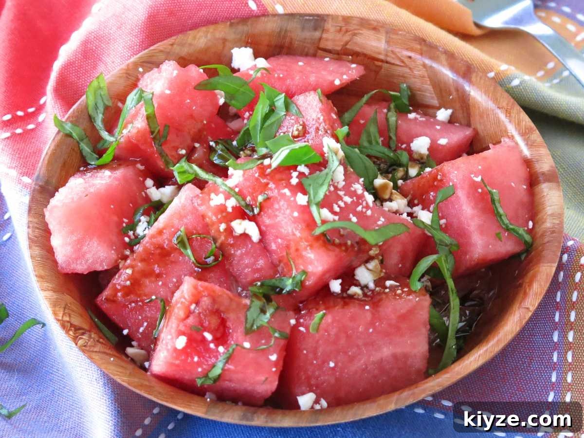 Chunks of watermelon topped with basil, balsamic glaze, and crumbled feta cheese in a brown bowl on a colorful napkin.