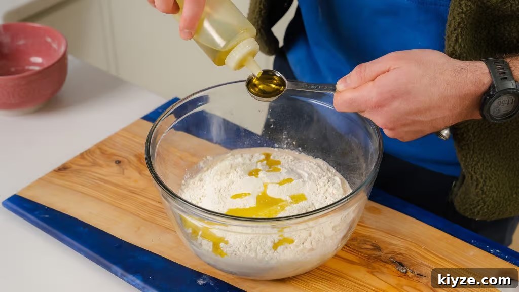 Homemade Pizza Dough Recipe for Crispy, Stretchy Crusts 3 The image shows the process of adding olive oil to a glass bowl filled with flour, one of the key ingredients for making homemade pizza dough. A person is holding a tablespoon while pouring olive oil from a squeeze bottle, ensuring the right amount is added to the mixture. The bowl rests on a wooden cutting board, and the scene captures a crucial step in combining the ingredients that will form a smooth and elastic dough, essential for creating a flavorful pizza crust.