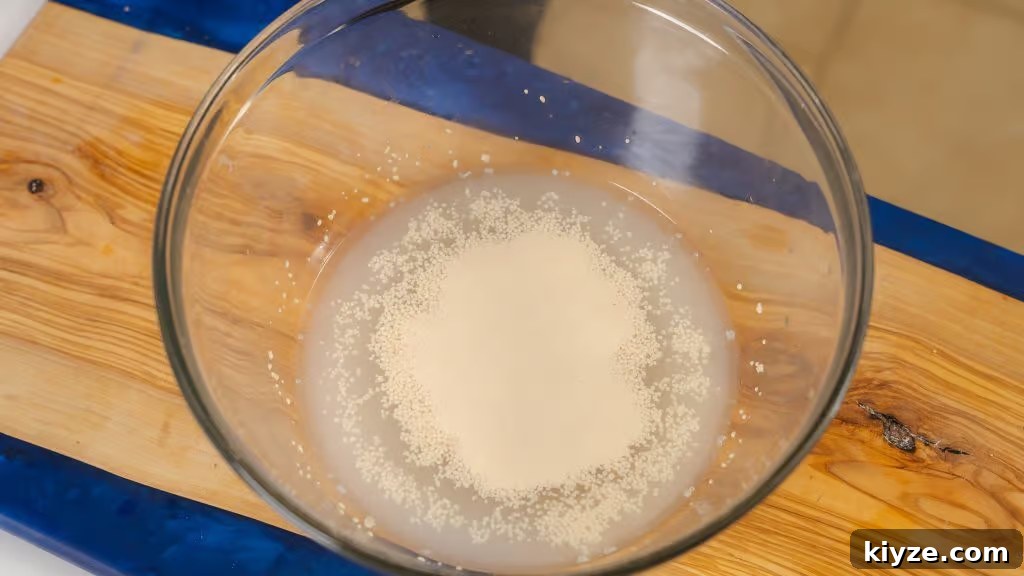 Homemade Pizza Dough Recipe for Crispy, Stretchy Crusts 4 The image shows a glass bowl containing warm water with yeast being activated, a crucial first step in making homemade pizza dough. The yeast is beginning to dissolve and foam, indicating it is alive and ready to leaven the dough. The bowl is placed on a wooden cutting board, setting the stage for the dough-making process. This step is essential for achieving a light and airy pizza crust, with the yeast ensuring the dough rises properly during proofing.