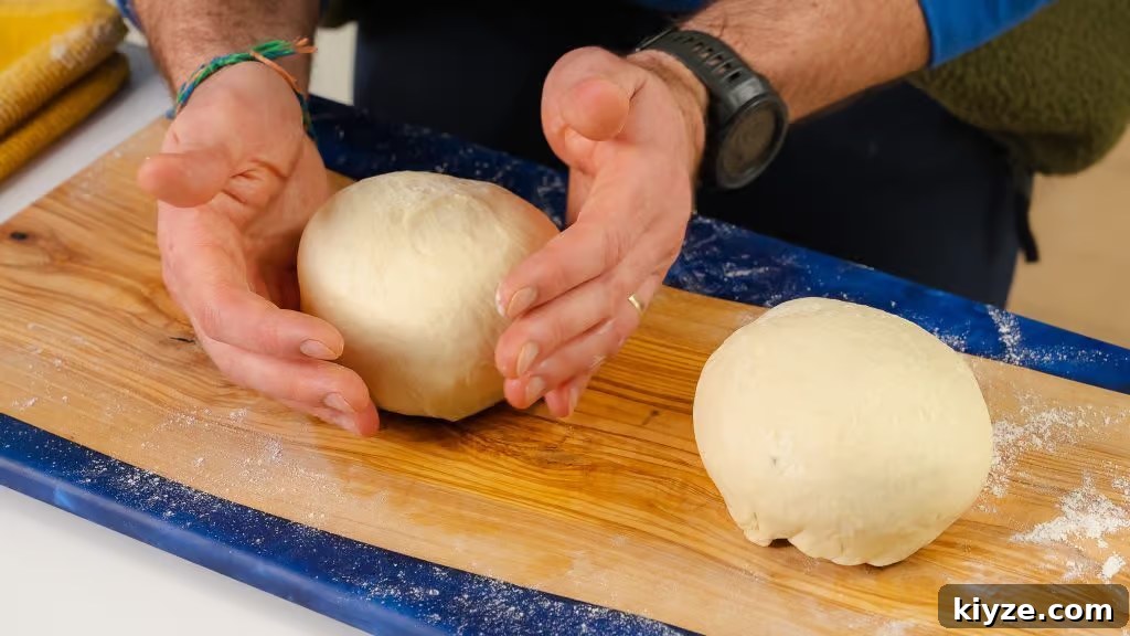 Homemade Pizza Dough Recipe for Crispy, Stretchy Crusts 5 The image shows two perfectly rounded balls of homemade pizza dough resting on a flour-dusted wooden board. A person’s hands gently shape one of the dough balls, ensuring a smooth and even surface, ready for proofing. The scene emphasizes the careful preparation process that goes into making pizza dough from scratch, highlighting the dough's soft and elastic texture, ideal for creating a perfect pizza crust.