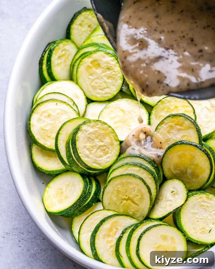 creamy sauce being added over the sliced zucchini in a white dish