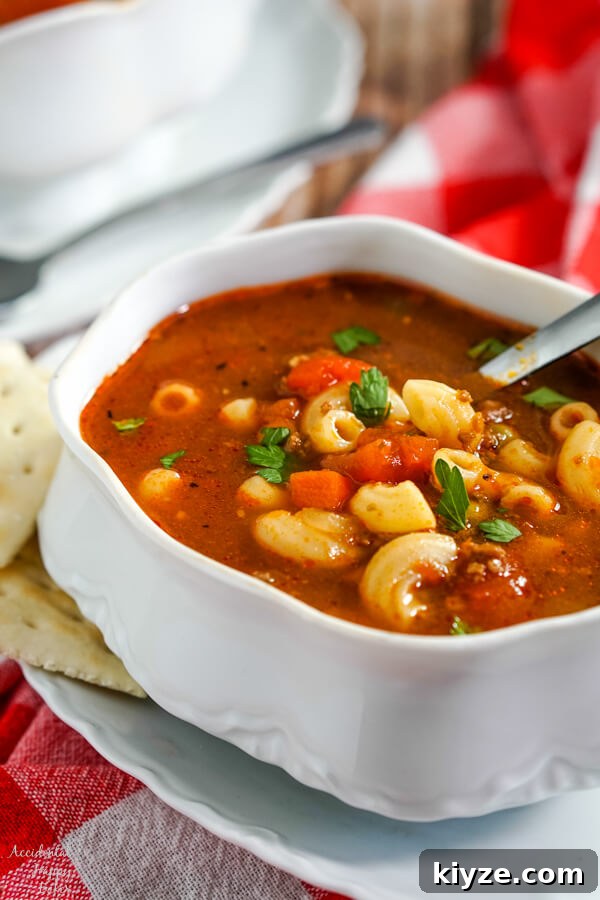A white soup tureen full of hamburger macaroni soup with a soup spoon in the bowl and crackers to the side.