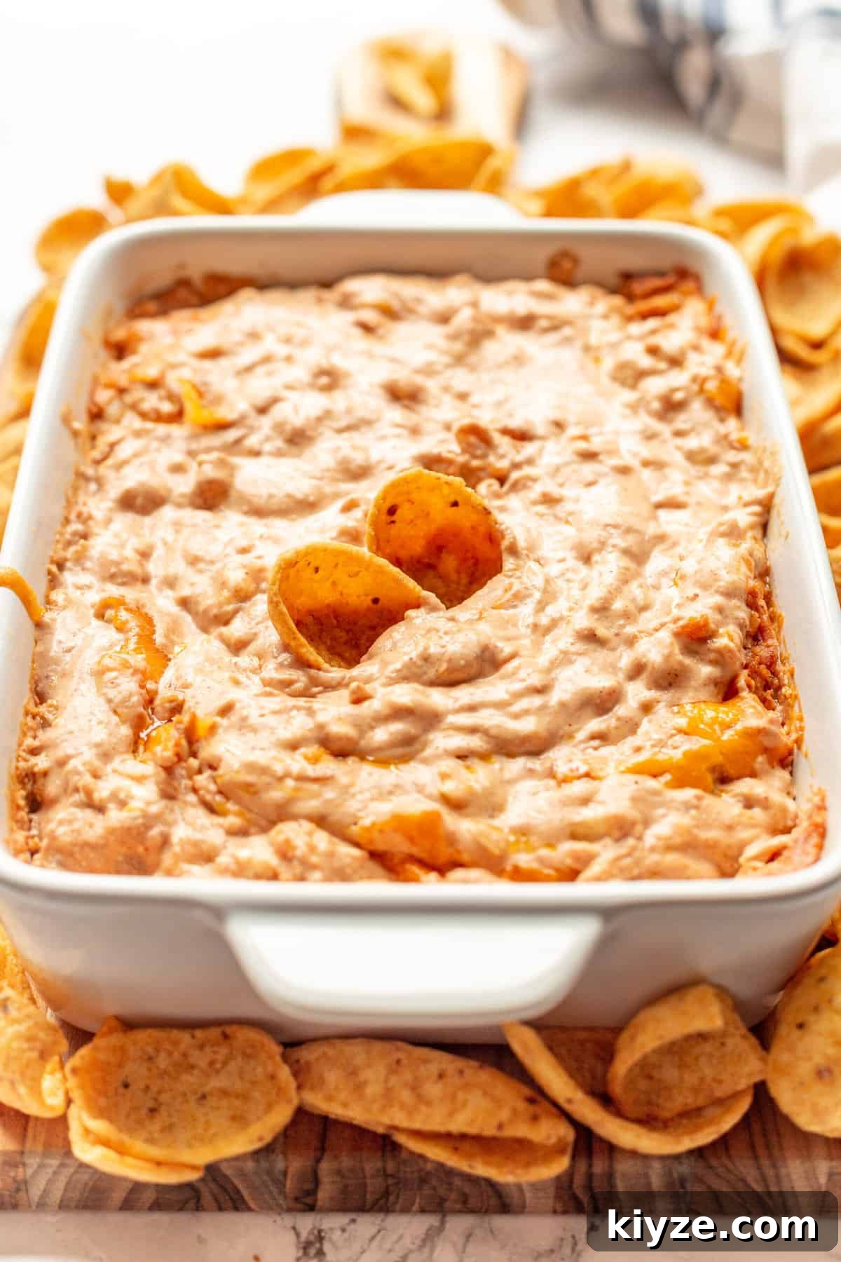 Creamy Cheesy Refried Bean Dip for Parties and Game Day 2 Cheesy refried bean dip with two fritos sitting in a large white casserole dish surrounded by frito chips.