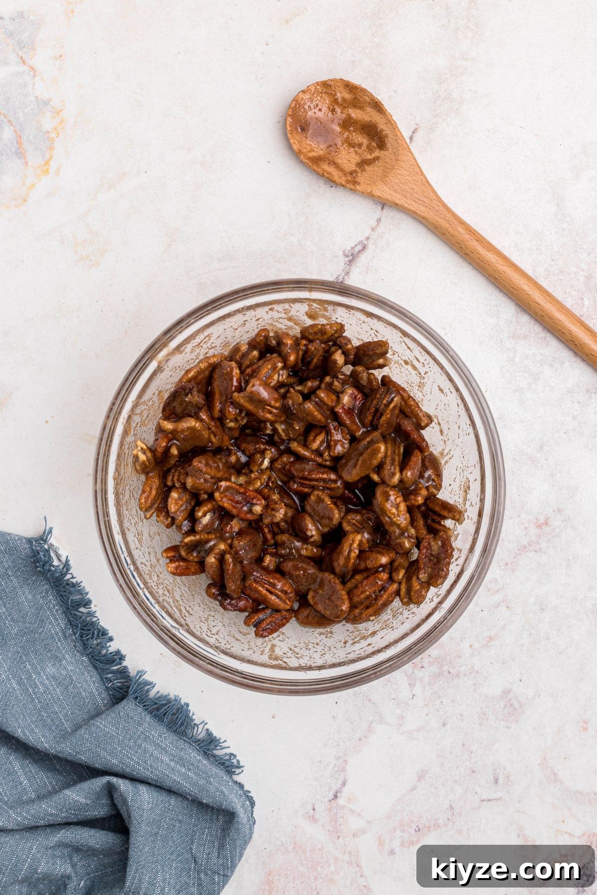 Cinnamon Sugar Candied Pecans Recipe for Snacking & Gifts 7 Sugars and cinnamon being tossed with coated pecans in a clear glass bowl on a marble table.