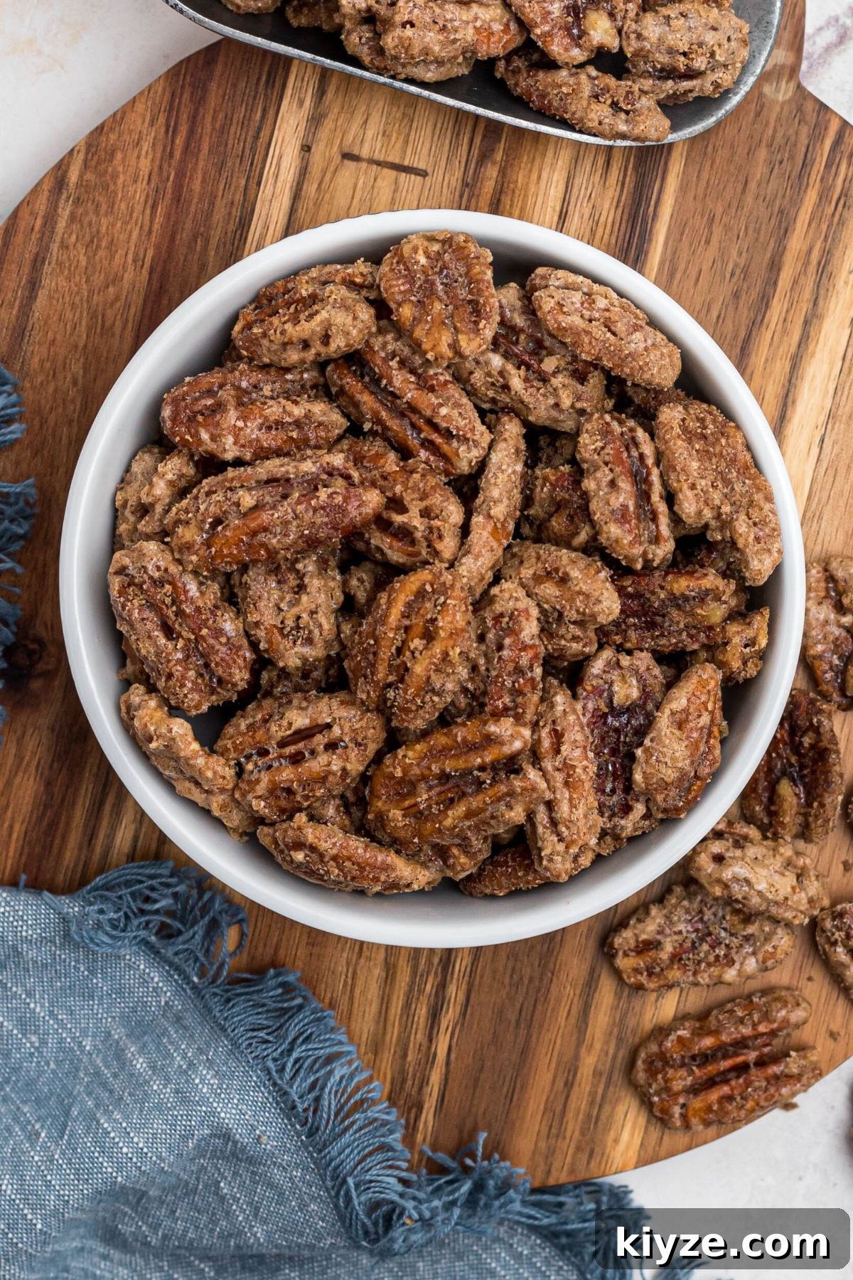 Cinnamon Sugar Candied Pecans Recipe for Snacking & Gifts 10 Candied pecans in a small white bowl, with a blue linen and scattered pecans nearby.