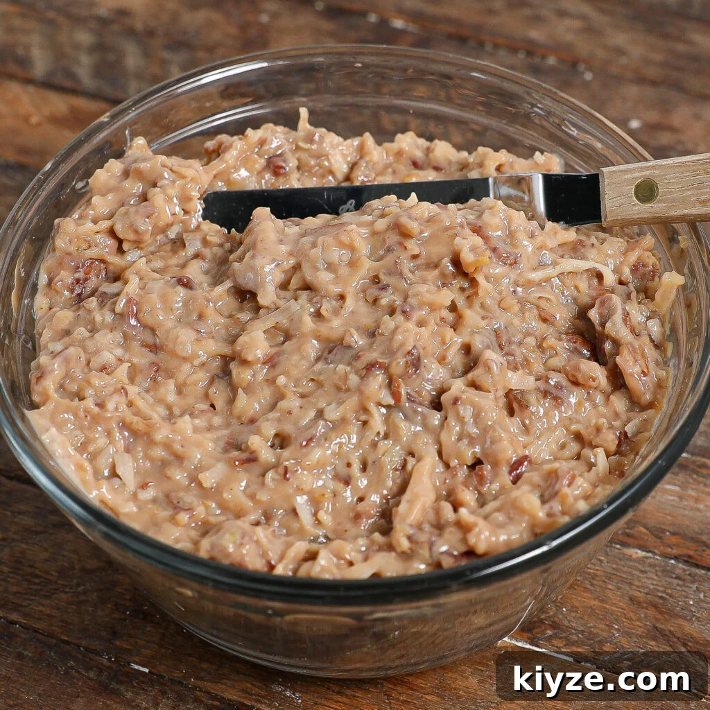 German Chocolate Cake Frosting Recipe: Rich Coconut-Pecan Icing 2 A glass bowl filled with thick, chunky German Chocolate Cake Frosting—packed with shredded coconut and chopped pecans—sits on a wooden surface with a knife resting on the bowl's edge.