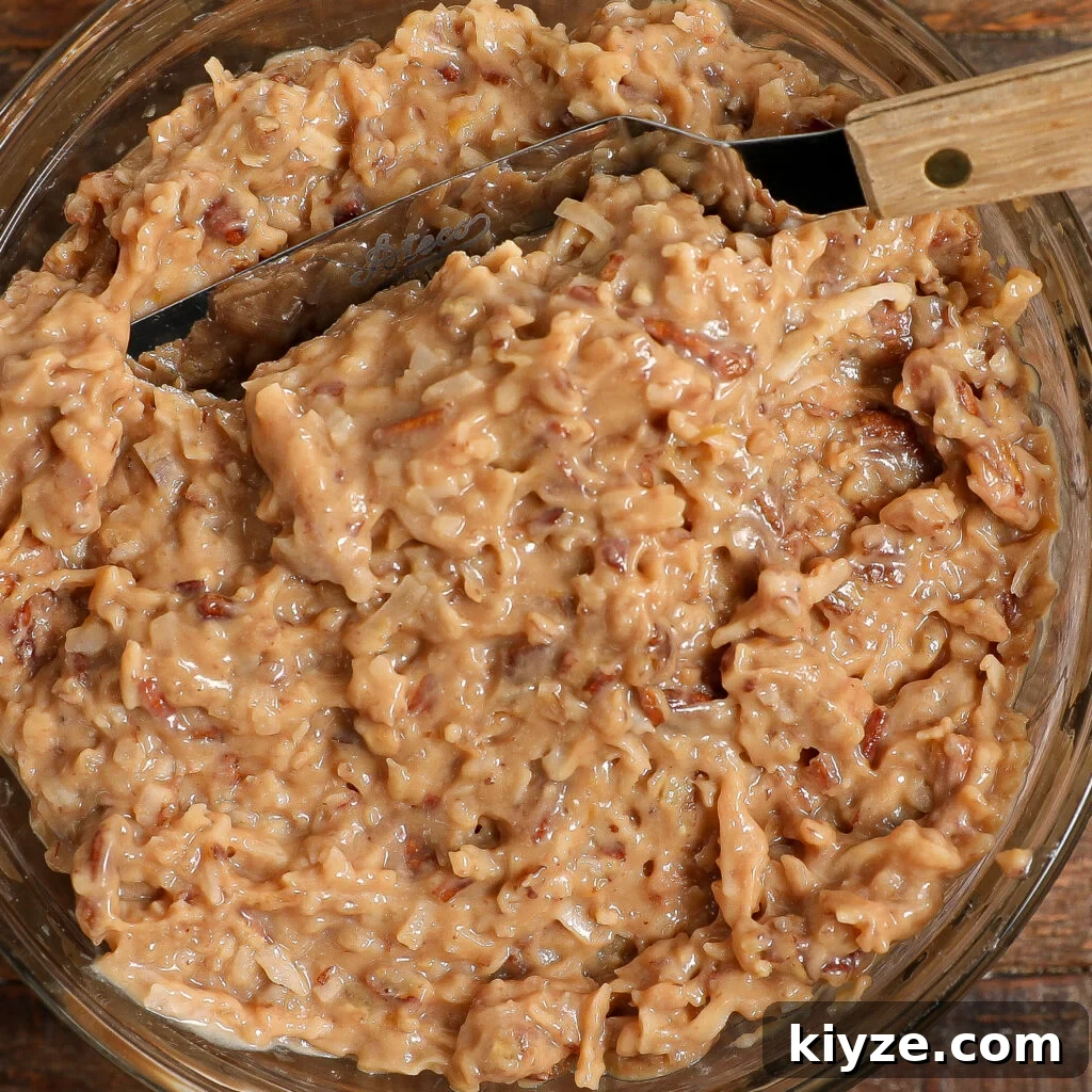German Chocolate Cake Frosting Recipe: Rich Coconut-Pecan Icing 3 A close-up of a bowl filled with thick, creamy German Chocolate Cake Frosting packed with shredded coconut and chopped pecans, with a metal spatula resting inside.