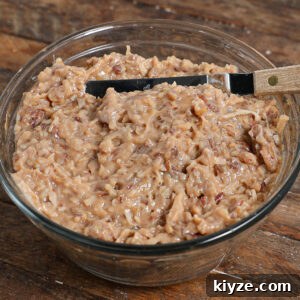 German Chocolate Cake Frosting Recipe: Rich Coconut-Pecan Icing 8 A glass bowl filled with thick, chunky German Chocolate Cake Frosting—packed with shredded coconut and chopped pecans—sits on a wooden surface with a knife resting on the bowl's edge.