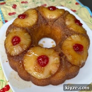 A pineapple upside-down bundt cake topped with pineapple rings and red maraschino cherries, displayed on a white plate over a yellow cloth with red cherry patterns.