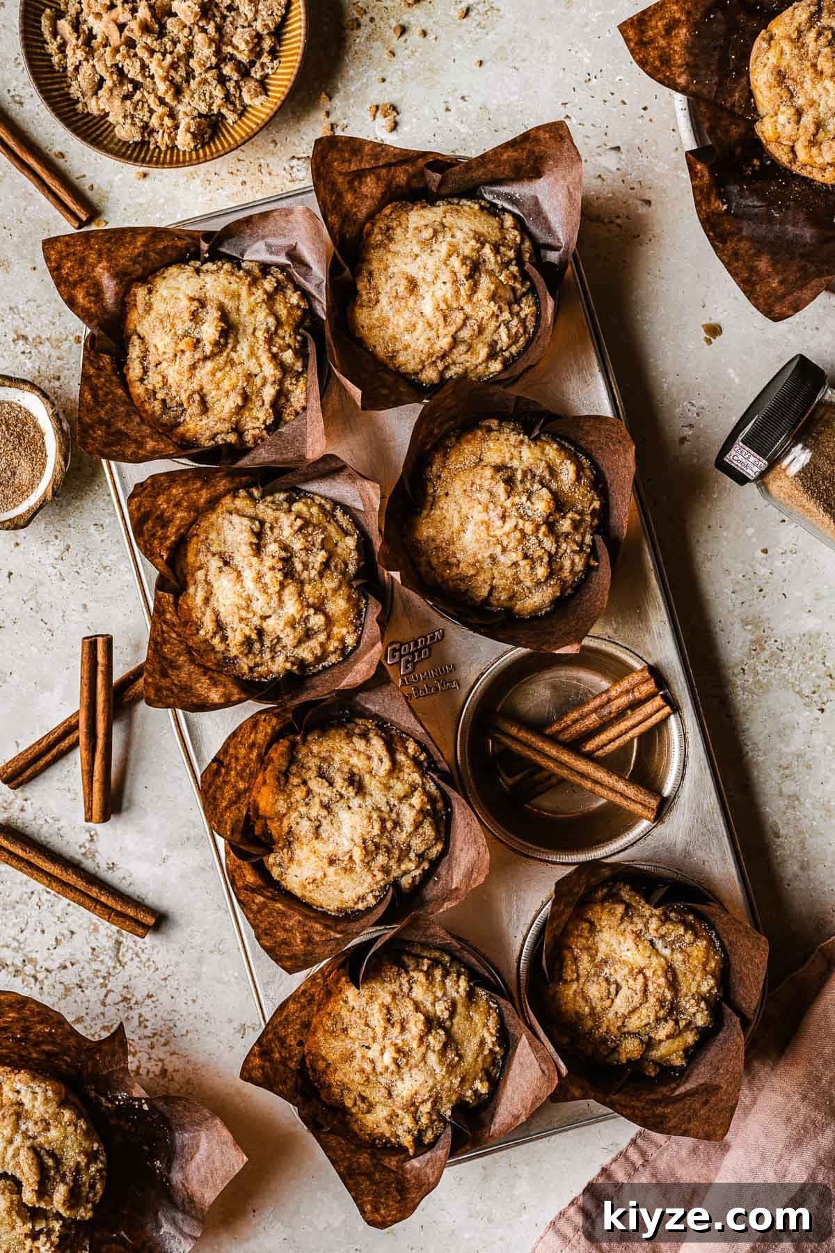 Cinnamon Streusel Muffins Recipe with Crunchy Brown Sugar Topping 2 Streusel topped muffins in brown paper parchment liners in a muffin tin.