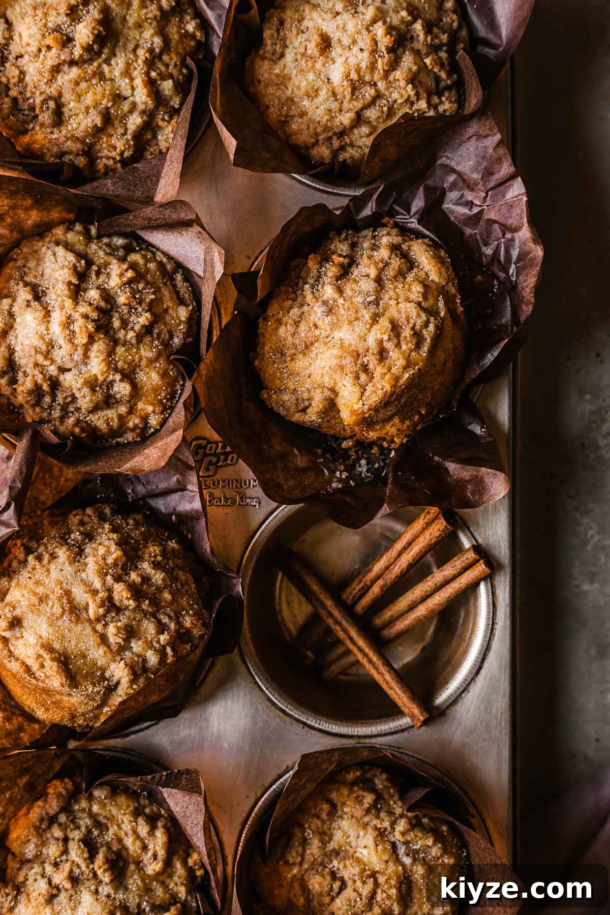 Cinnamon Streusel Muffins Recipe with Crunchy Brown Sugar Topping 11 Streusel topped muffins in brown parchment wrappers in a muffin tin. One hole of the muffin tin has three sticks of cinnamon inside.