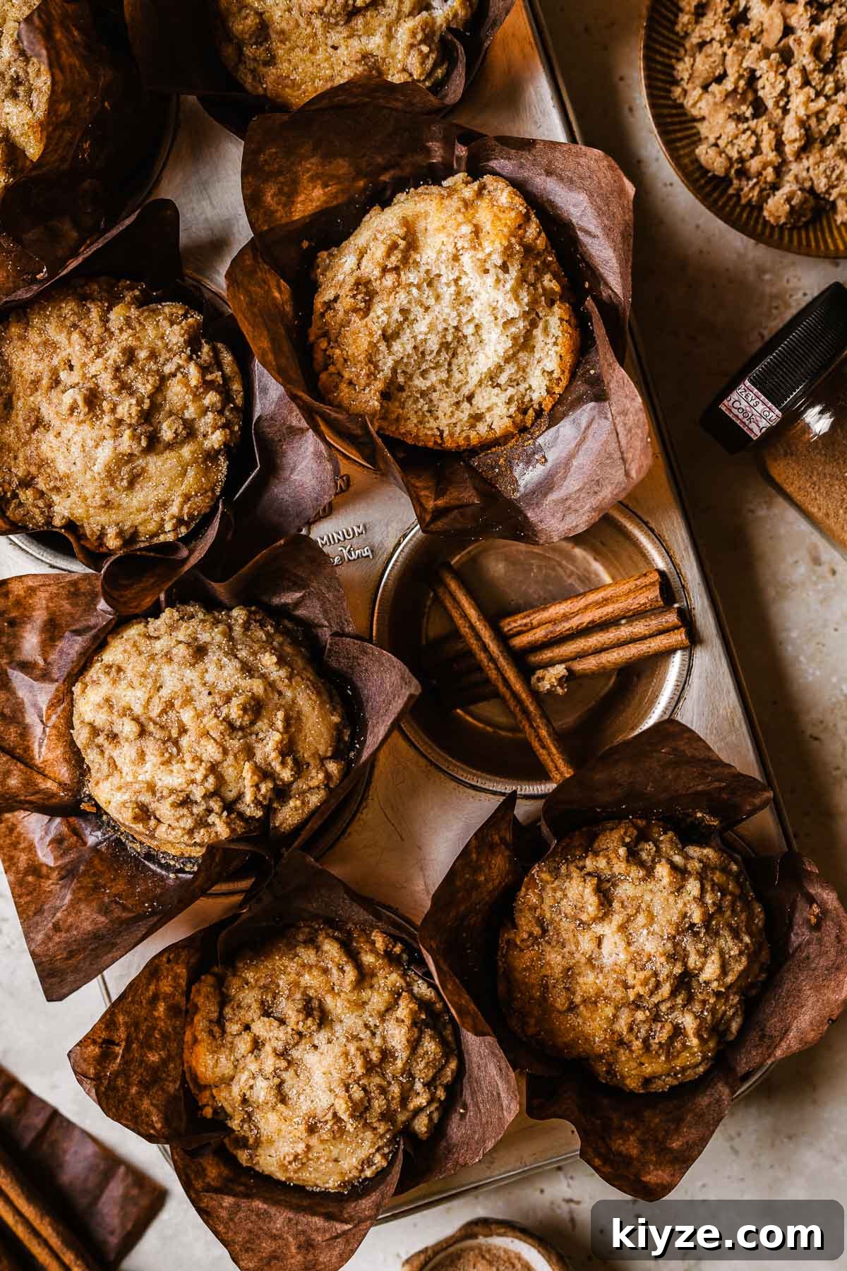 Cinnamon Streusel Muffins Recipe with Crunchy Brown Sugar Topping 13 Cinnamon streusel muffins in brown parchment paper wrappers. One muffin has been broken open to show the crumb inside.