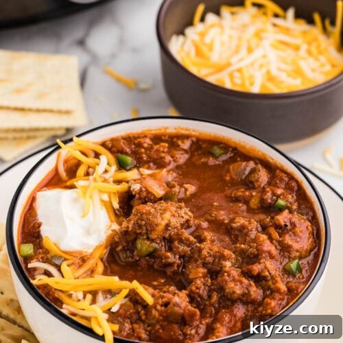 Slow Cooker No-Bean Chili Recipe for Tender, Flavorful Meat 11 dinner plate with bowl of chili with sour cream and cheese on top, crackers on side, with bowl of cheese, crackers and black slow cooker in background