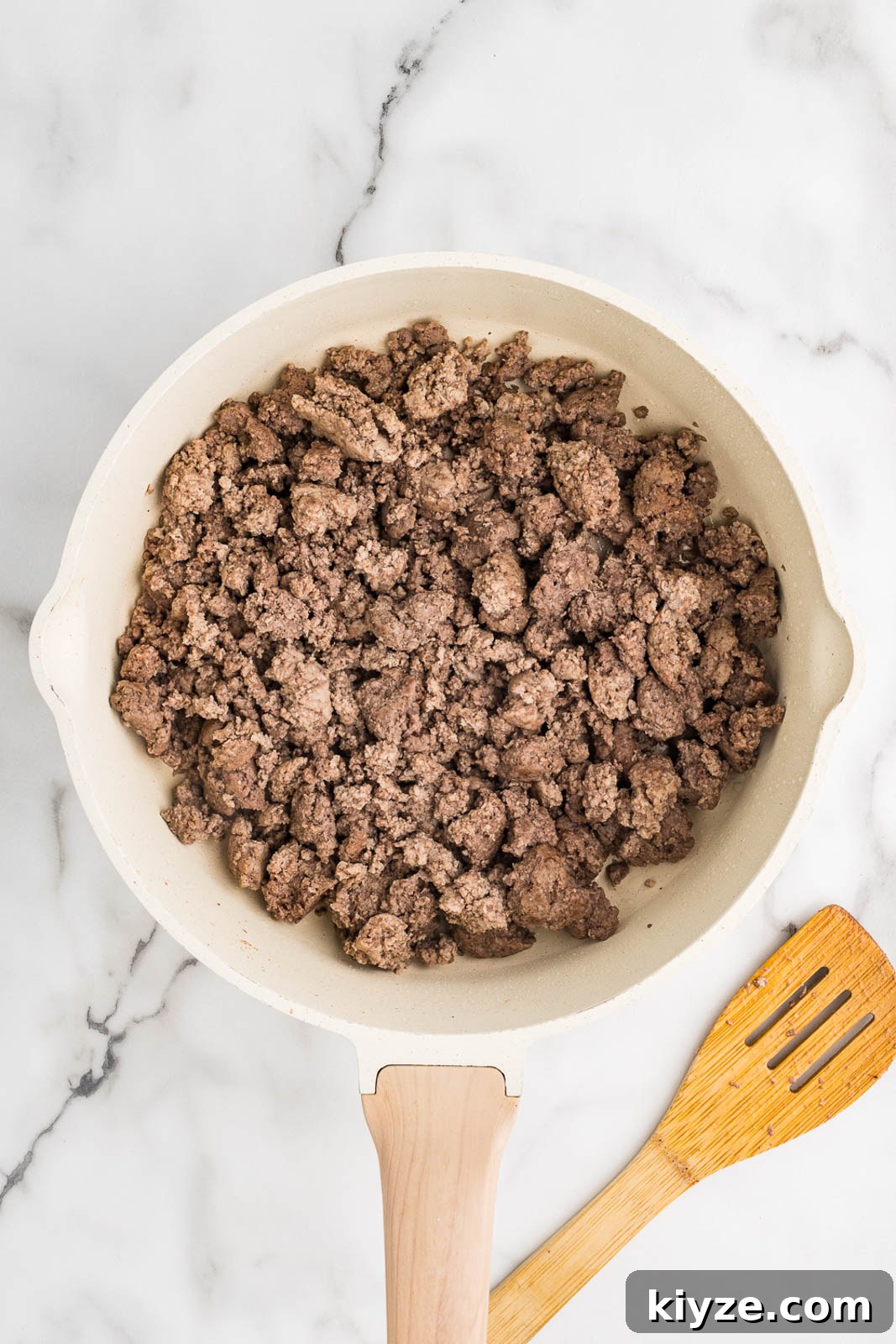Slow Cooker No-Bean Chili Recipe for Tender, Flavorful Meat 4 white skillet with wooden handle containing ground meats with wooden spoon on side
