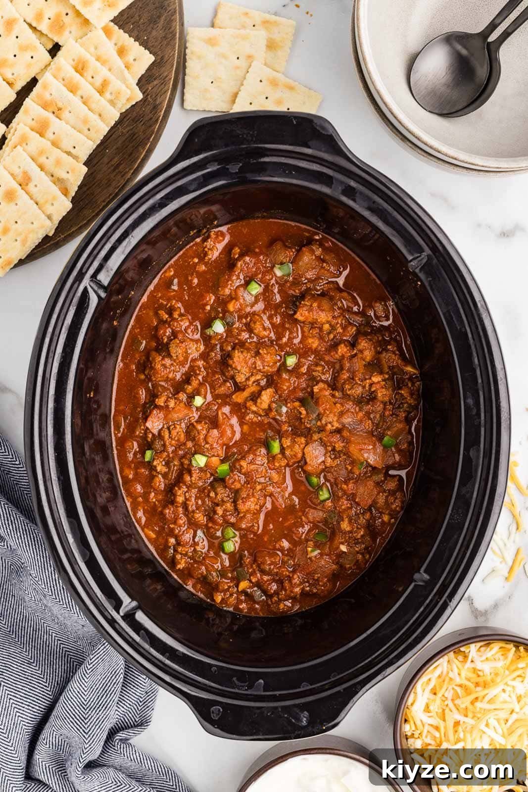Slow Cooker No-Bean Chili Recipe for Tender, Flavorful Meat 8 slow cooked chili recipe in black slow cooker, surrounded by crackers, bowls with spoons, cheese and sour cream