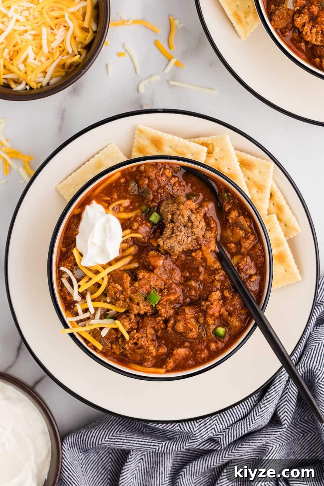 Slow Cooker No-Bean Chili Recipe for Tender, Flavorful Meat 10 overhead view of white dinner plate with bowl of chili topped with cheese and sour cream, crackers on the side, with bowls of cheese and sour cream and another bowl of chili in background