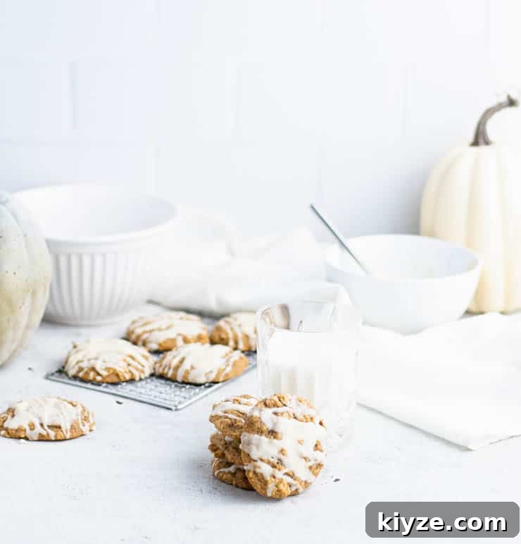 a stack of iced pumpkin cookies next to a short glass of milk with additional cookies, bowls and white and green pumpkins in the background against a tiled backdrop.