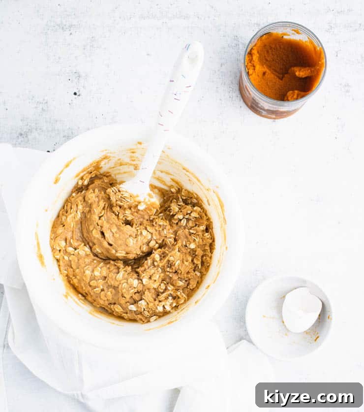 pumpkin cookie batter with visible bits of rolled oats in a white bowl next to a bowl with egg shells and a container of pumpkin puree.