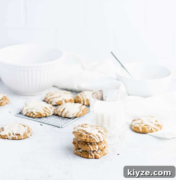 Iced pumpkin oatmeal cookies stacked in front of a cooling rack of cookies, and white bowls.