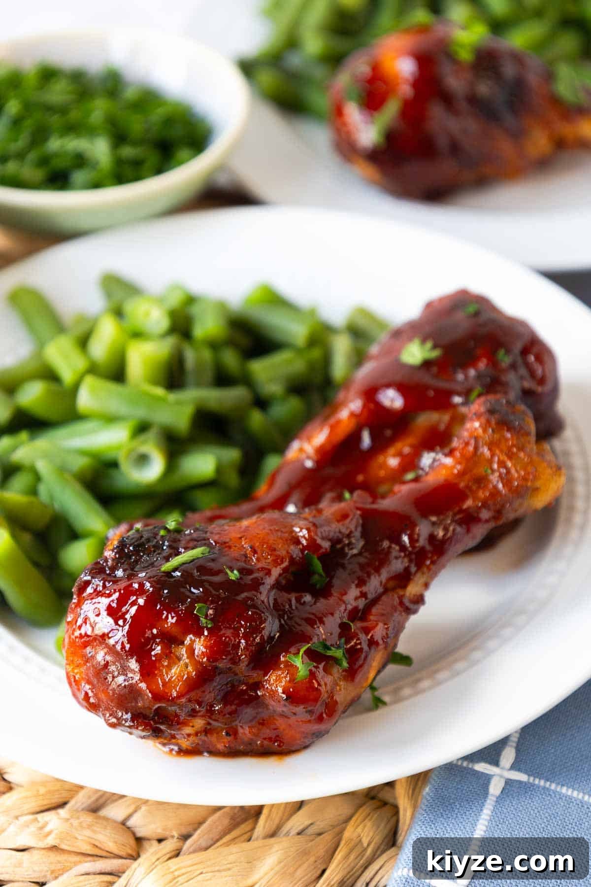 Crispy Oven-Baked BBQ Chicken Drumsticks 2 A baked barbecue chicken drumstick coated in thick glossy sauce sits on a plate with a side of chopped green beans. The sauce appears sticky and caramelized, with small flecks of herbs on top.