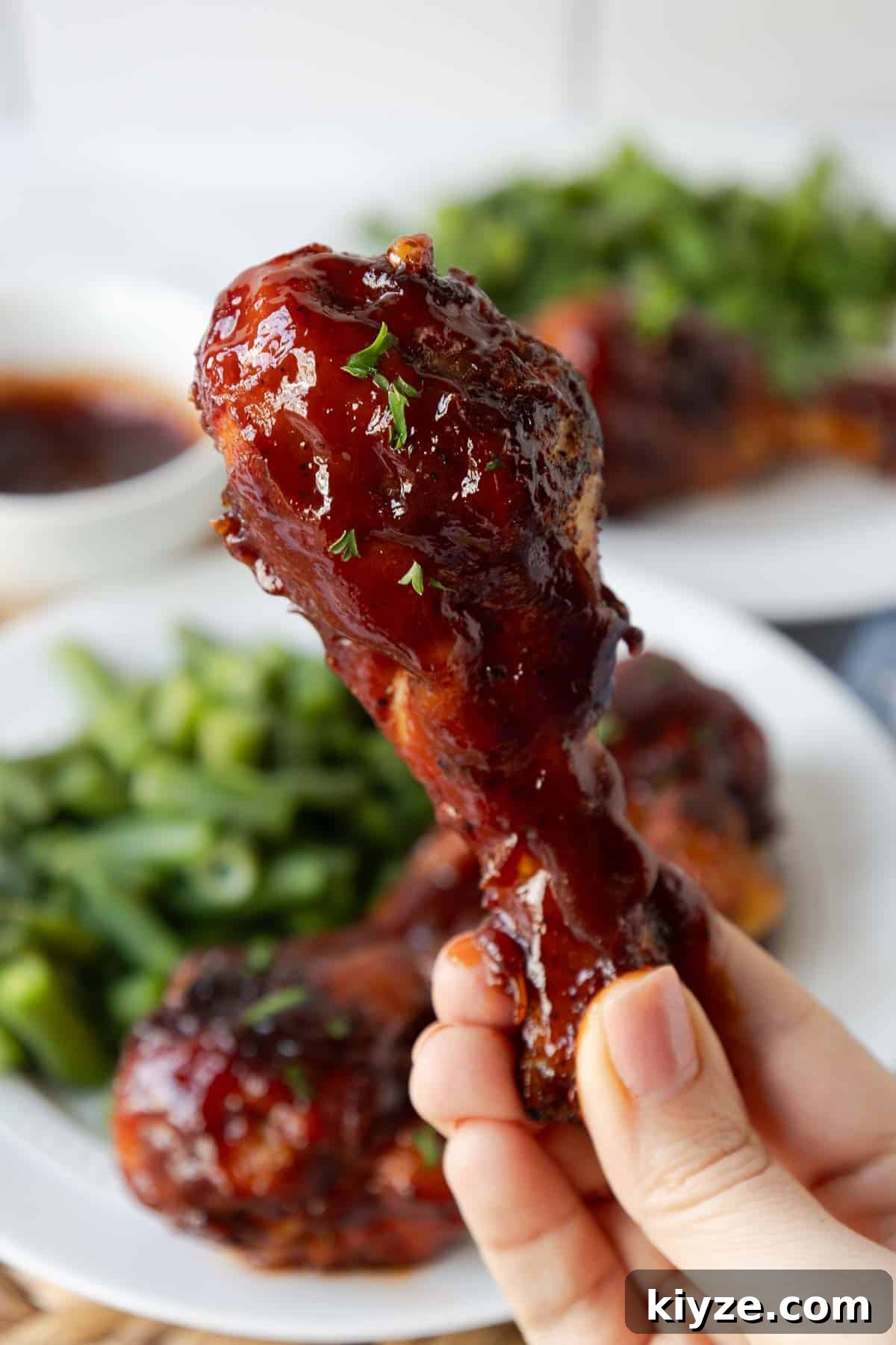 Crispy Oven-Baked BBQ Chicken Drumsticks 11 A hand holds a saucy baked chicken drumstick up close, showing the thick barbecue coating and tender texture. More glazed drumsticks and green beans are blurred in the background.