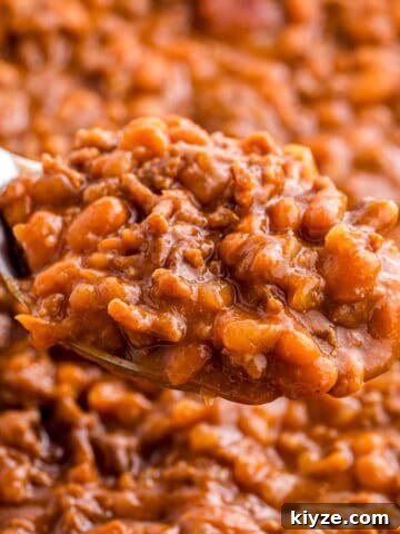 Spoonful of baked beans with ground beef held above the dish to show the thick texture and rich sauce. Tender beans and crumbled beef are visible in the close up.