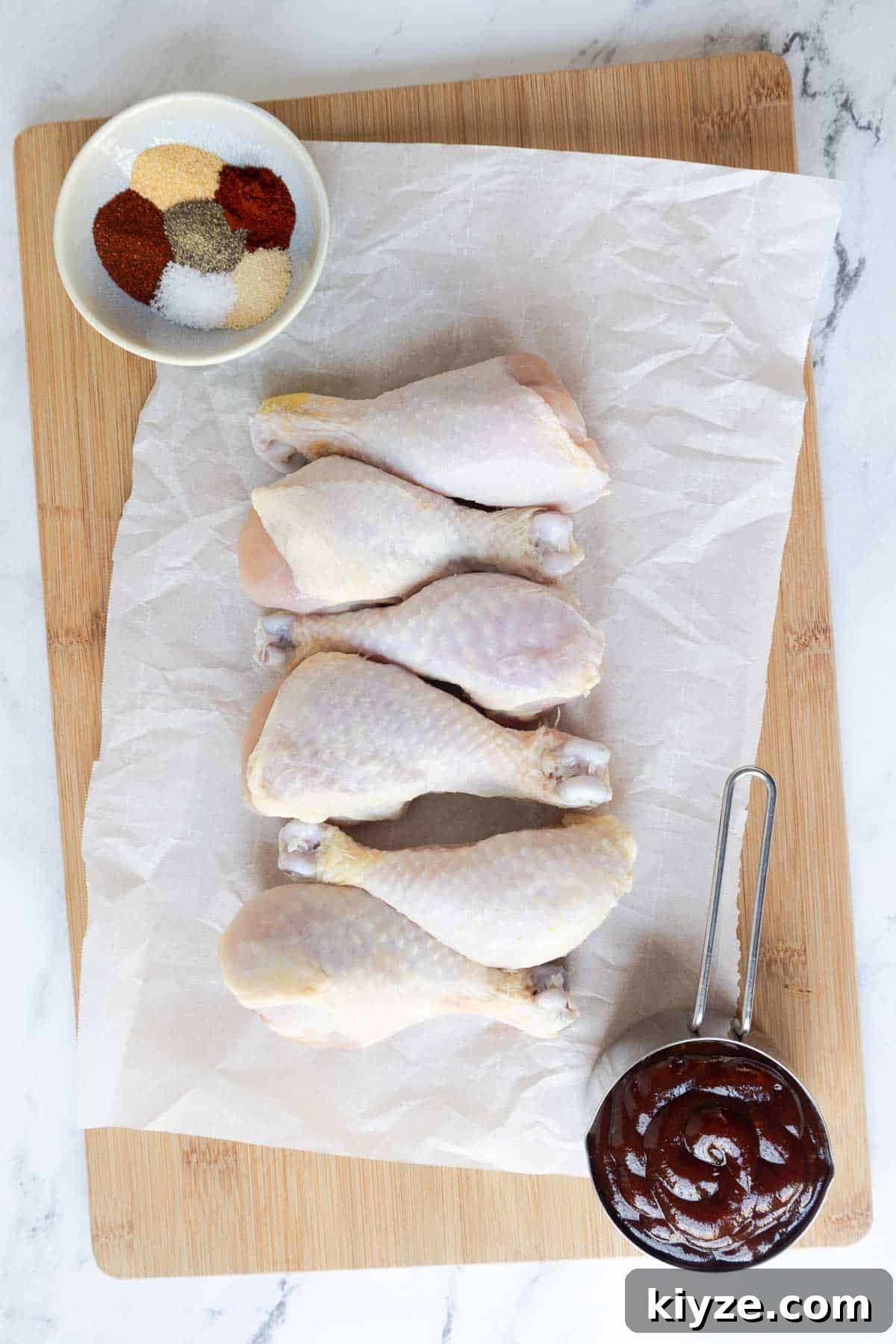 Crispy Oven-Baked BBQ Chicken Drumsticks 3 Raw chicken drumsticks are laid out on parchment paper next to a small bowl of mixed spices and a portion of barbecue sauce. The setup shows the ingredients before seasoning and cooking.