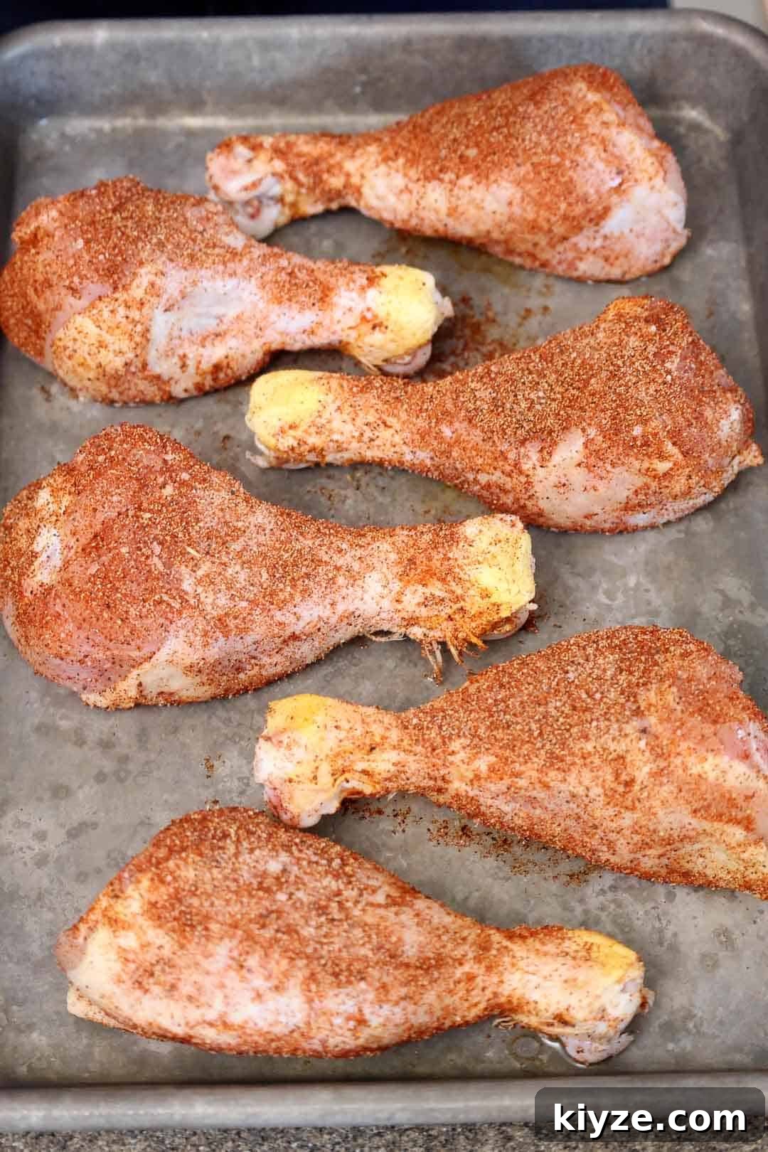 Crispy Oven-Baked BBQ Chicken Drumsticks 4 Chicken drumsticks coated in a dry spice rub sit on a baking tray before being cooked. The seasoning is evenly distributed across the skin, giving it a textured appearance.