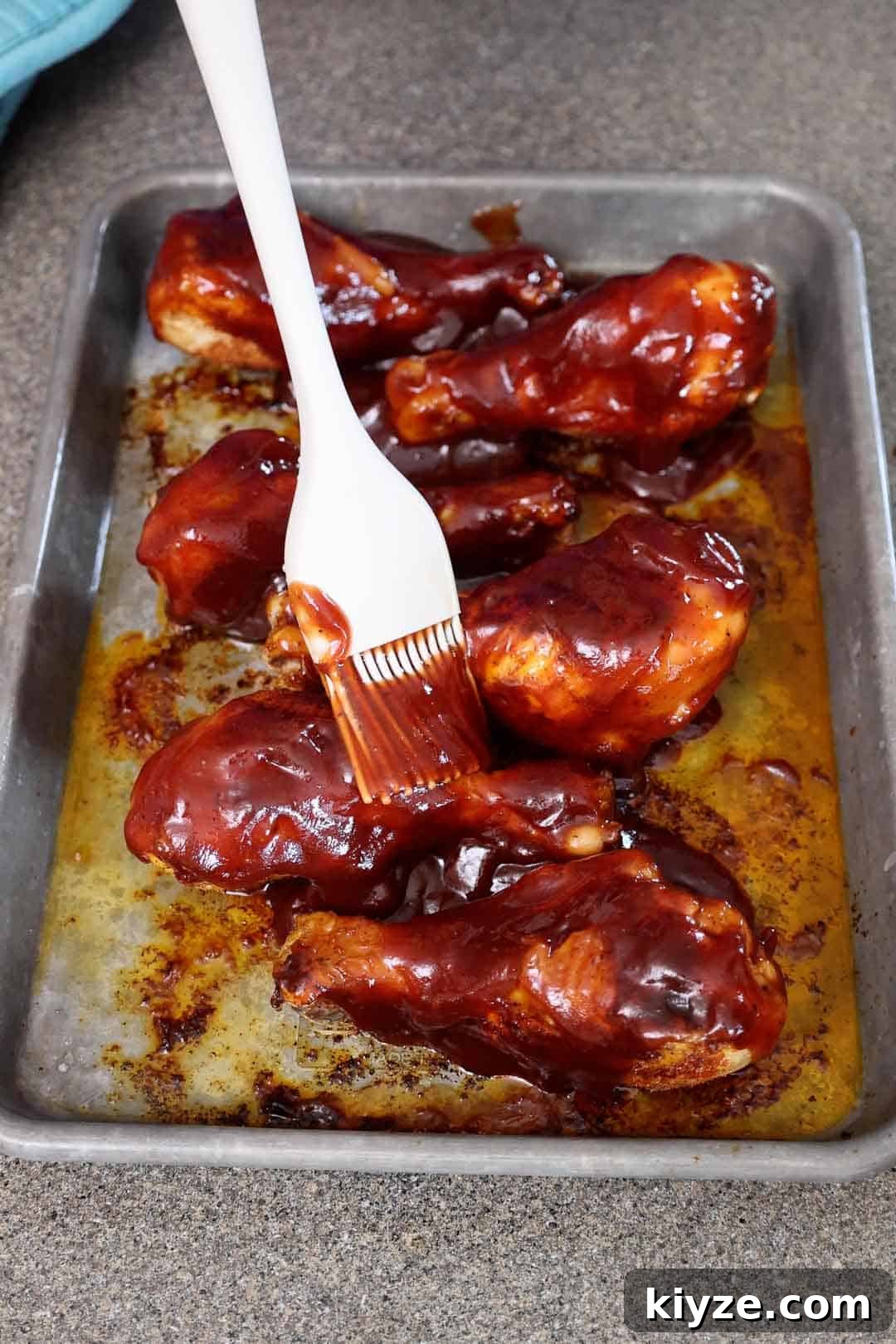 Crispy Oven-Baked BBQ Chicken Drumsticks 6 A brush spreads thick barbecue sauce over baked chicken drumsticks in a tray. The sauce is being applied generously, coating each piece before finishing in the oven.