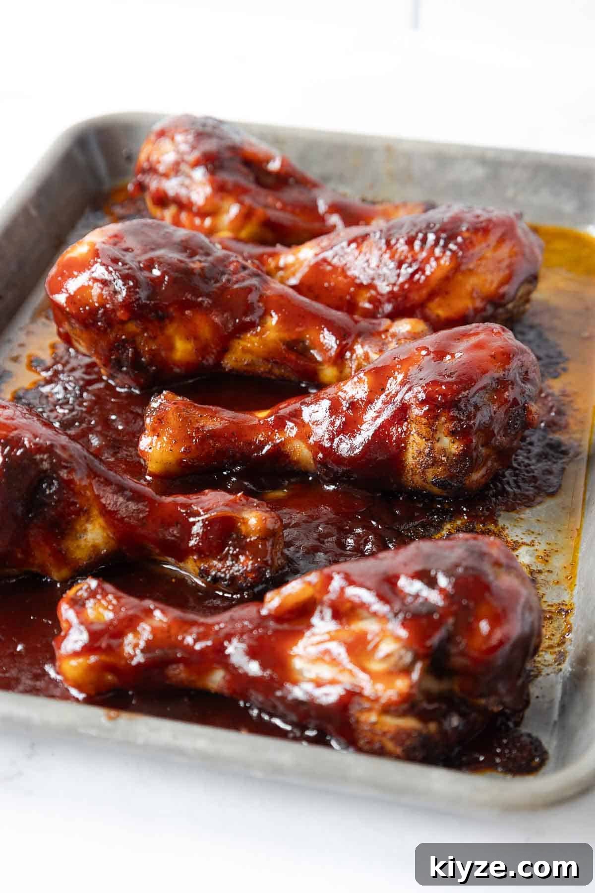 Crispy Oven-Baked BBQ Chicken Drumsticks 7 Fully cooked chicken drumsticks are coated in a glossy barbecue glaze and rest in a baking tray with caramelized sauce around them. The surface shows a mix of sticky glaze and lightly charred spots.