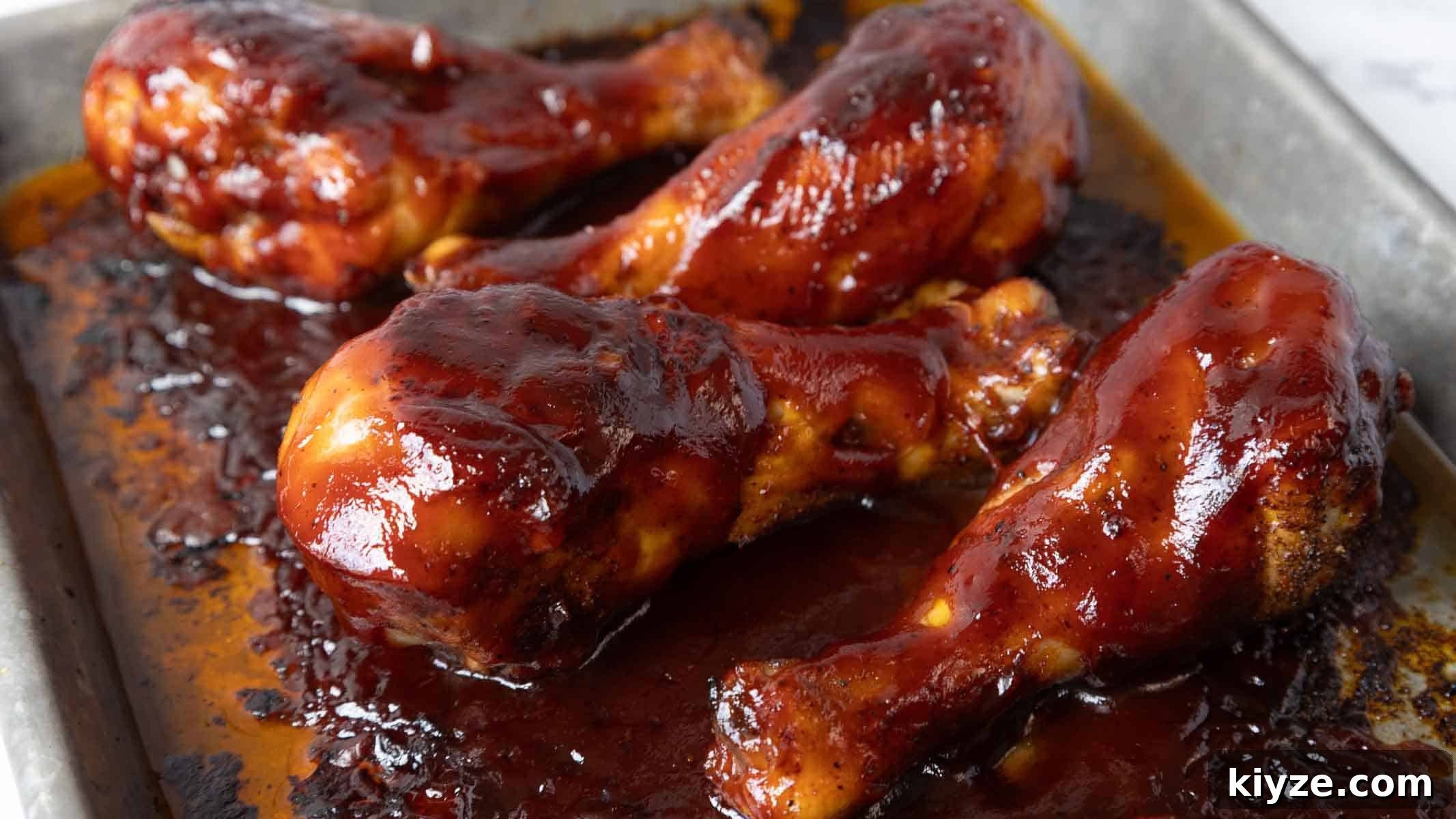 Crispy Oven-Baked BBQ Chicken Drumsticks 8 Chicken drumsticks rest in a baking tray coated in a layer of barbecue sauce that has thickened and caramelized during cooking. The sauce pools around the drumsticks with darker baked edges.