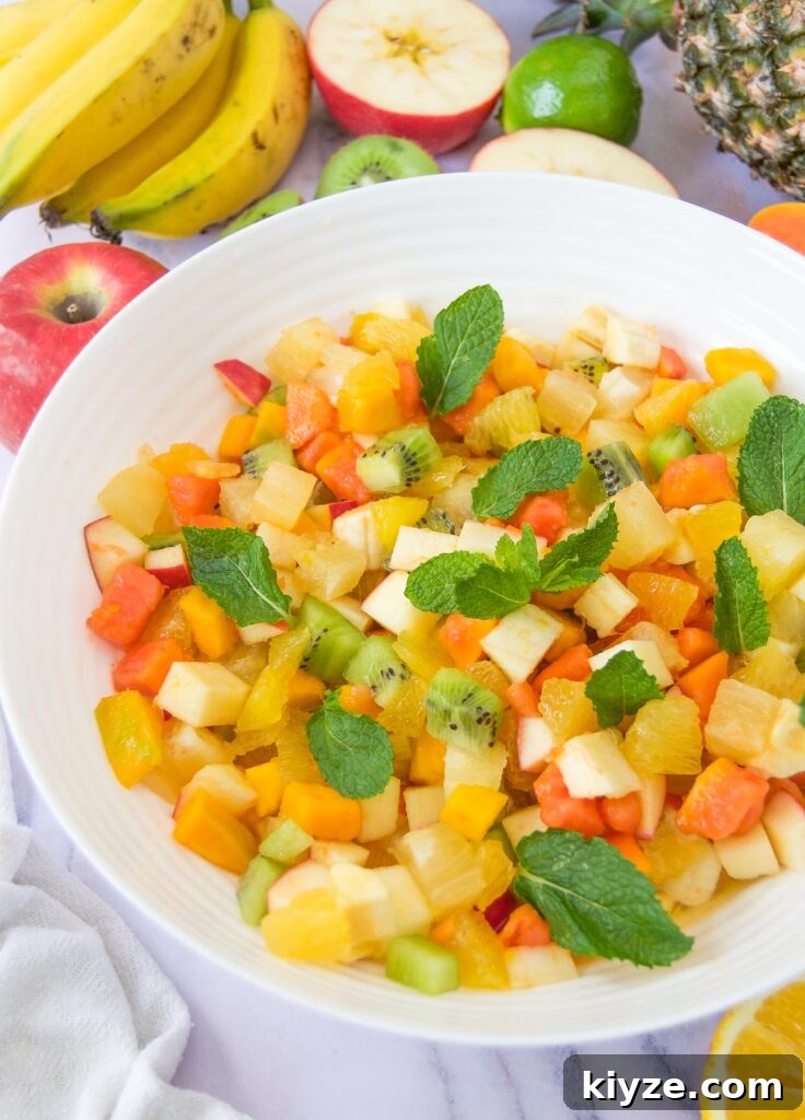 Tropical Fruit Salad with Mango, Pineapple and Coconut 7 a white bowl of an easy fruit salad with tropical fruits garnished with mint leaves and surrounded by more fruits