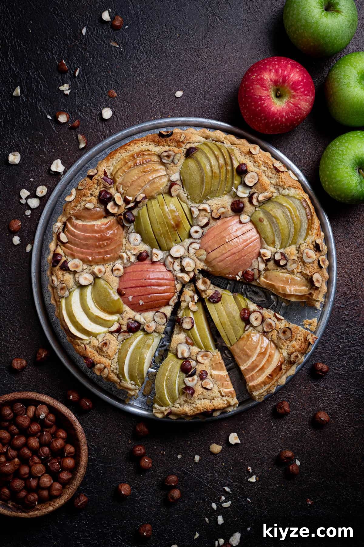 Vegan Apple Hazelnut Frangipane Tart 18 The sliced apple hazelnut frangipane tart on a dark surface with apples and a bowl of hazelnuts.