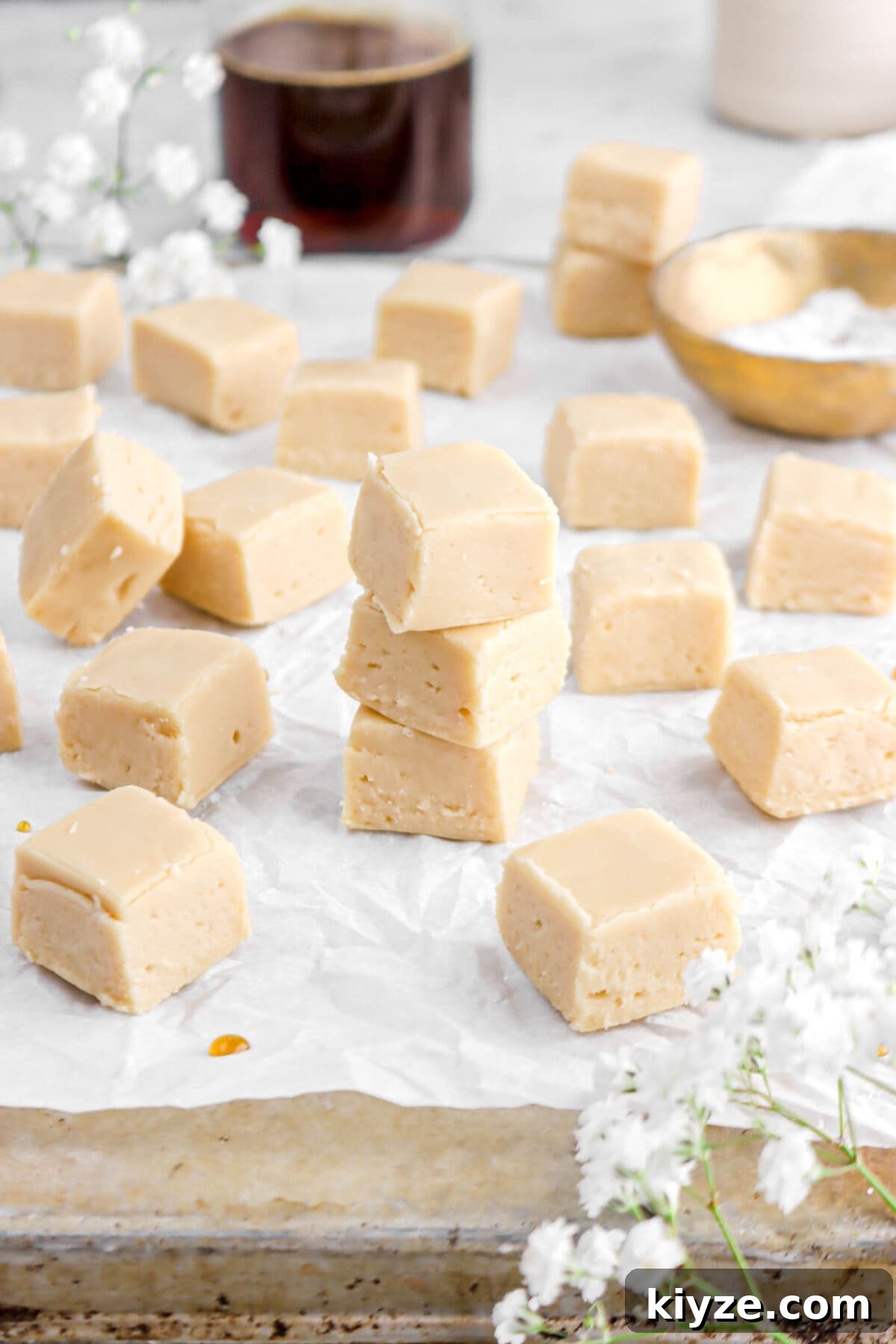 Old-Fashioned Maple Fudge Recipe 15 maple fudge on parchment paper on old sheet pan with white flowers around, gold bowl, and glass of maple syrup behind.