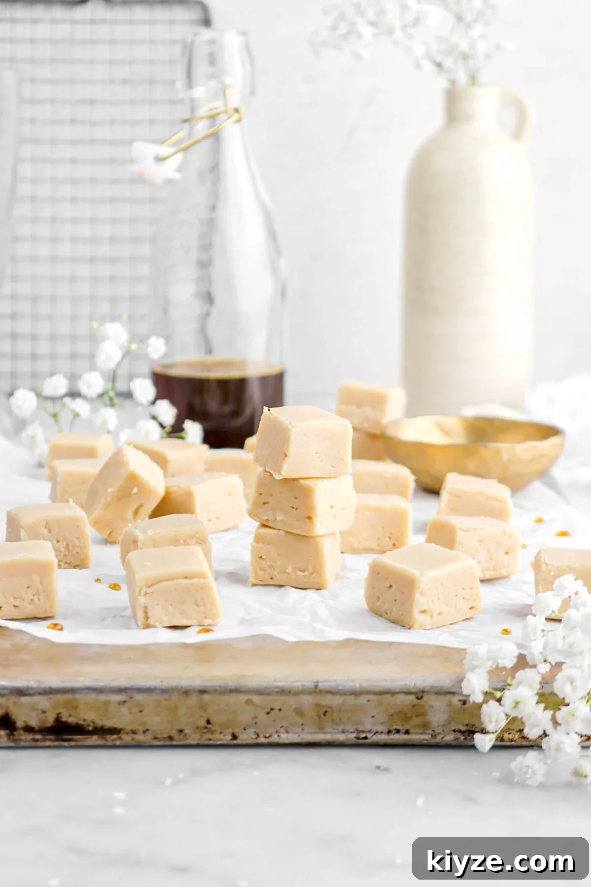 Old-Fashioned Maple Fudge Recipe 3 maple fudge on upside down sheet pan with gold bowl behind, glass of maple syrup, and vase behind on marble surface.