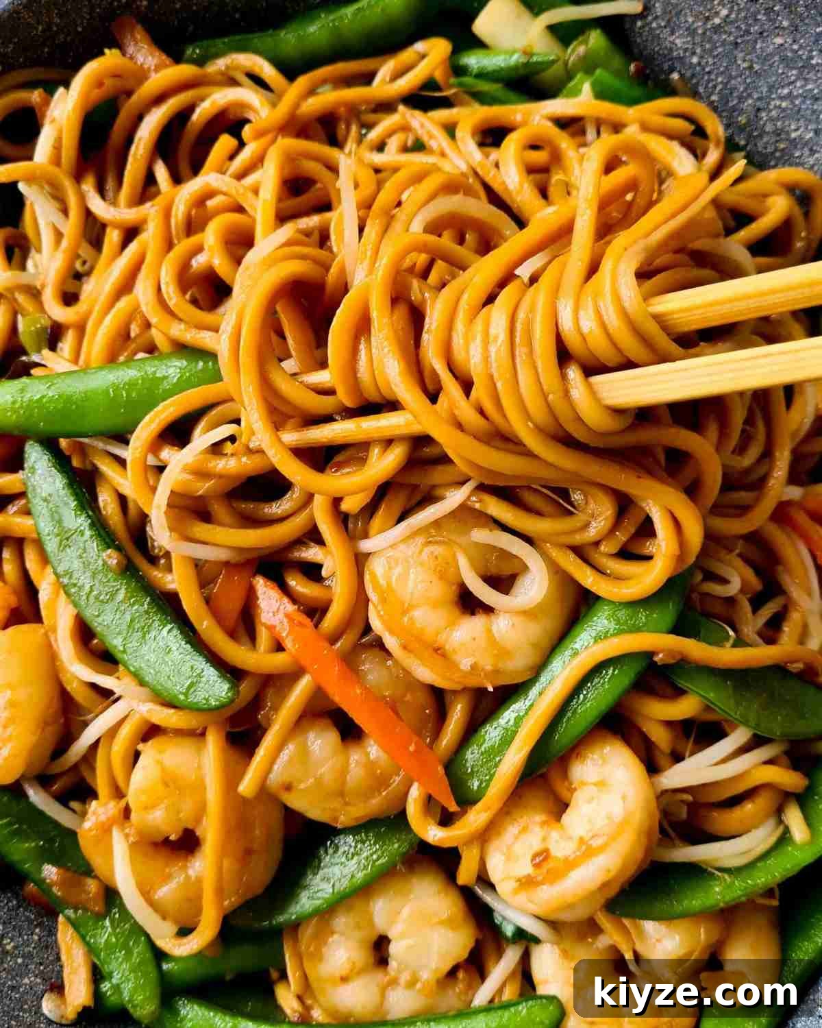 Wok Fried Prawn Noodles with Garlic and Chili 2 Hokkien noodles held up by a pair of chopsticks with prawns and vegetables in a grey fry pan