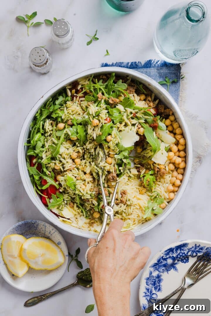 Fresh Pesto and Orzo Salad with Cherry Tomatoes and Basil 7 Pesto and orzo salad being tossed in a large mixing bowl by a woman's hand holding tongs.